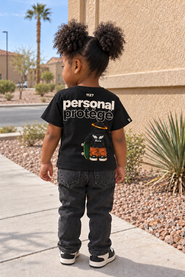 Personal Protégé Kids Tee — toddler back view beside a tan stucco wall